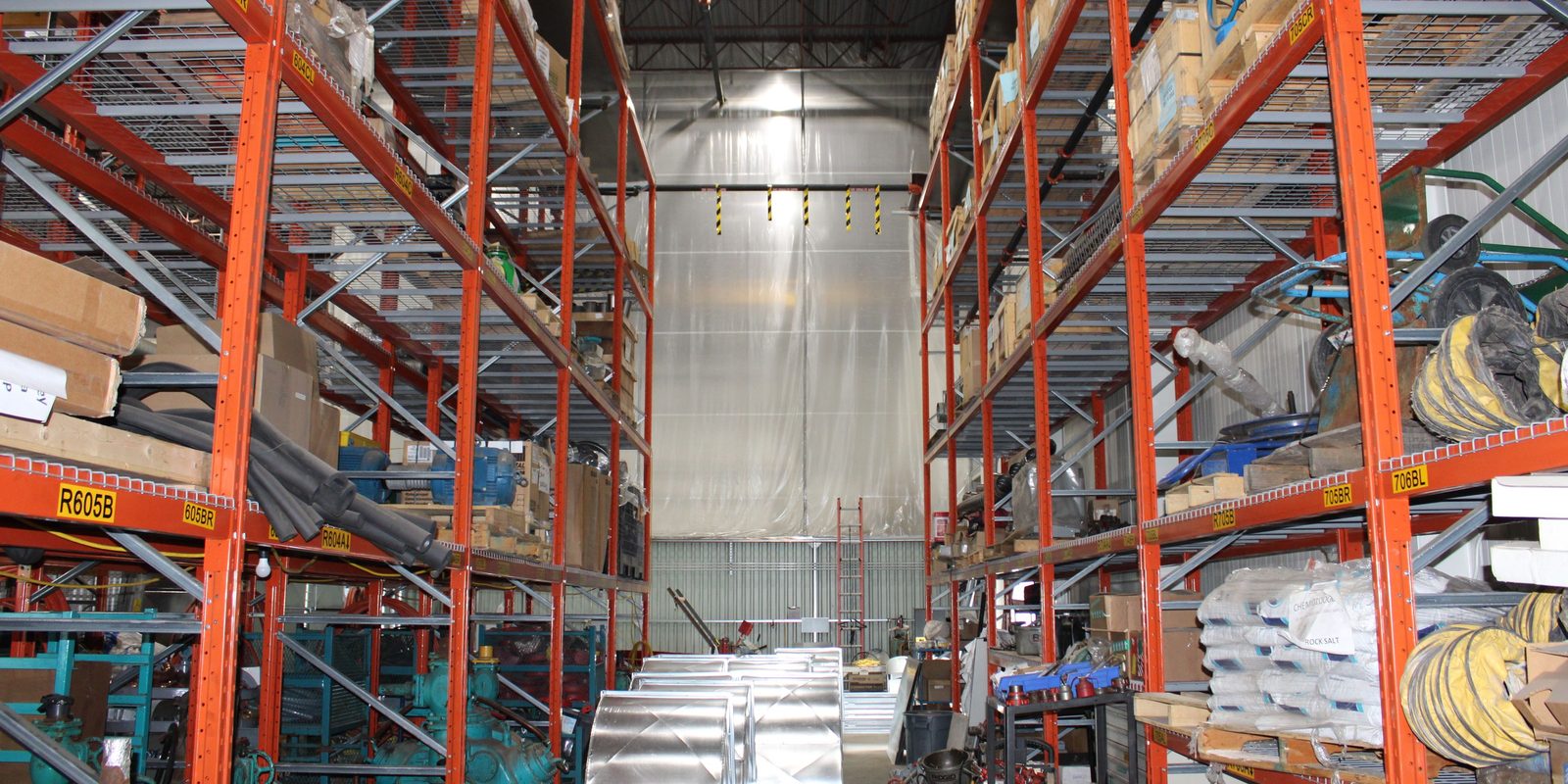 Tall orange pallet racks stocked with boxed parts, ductwork and coiled hose on both sides of a bright warehouse aisle in Burlington, Ontario