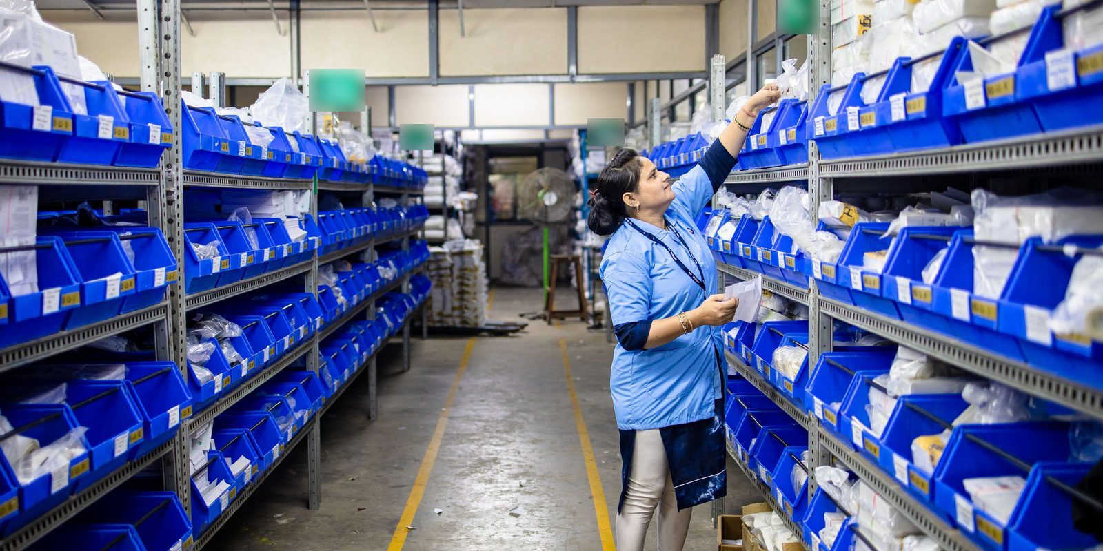 A worker in a light blue uniform takes a part from labelled blue bins on long steel shelves in a bright, orderly parts store