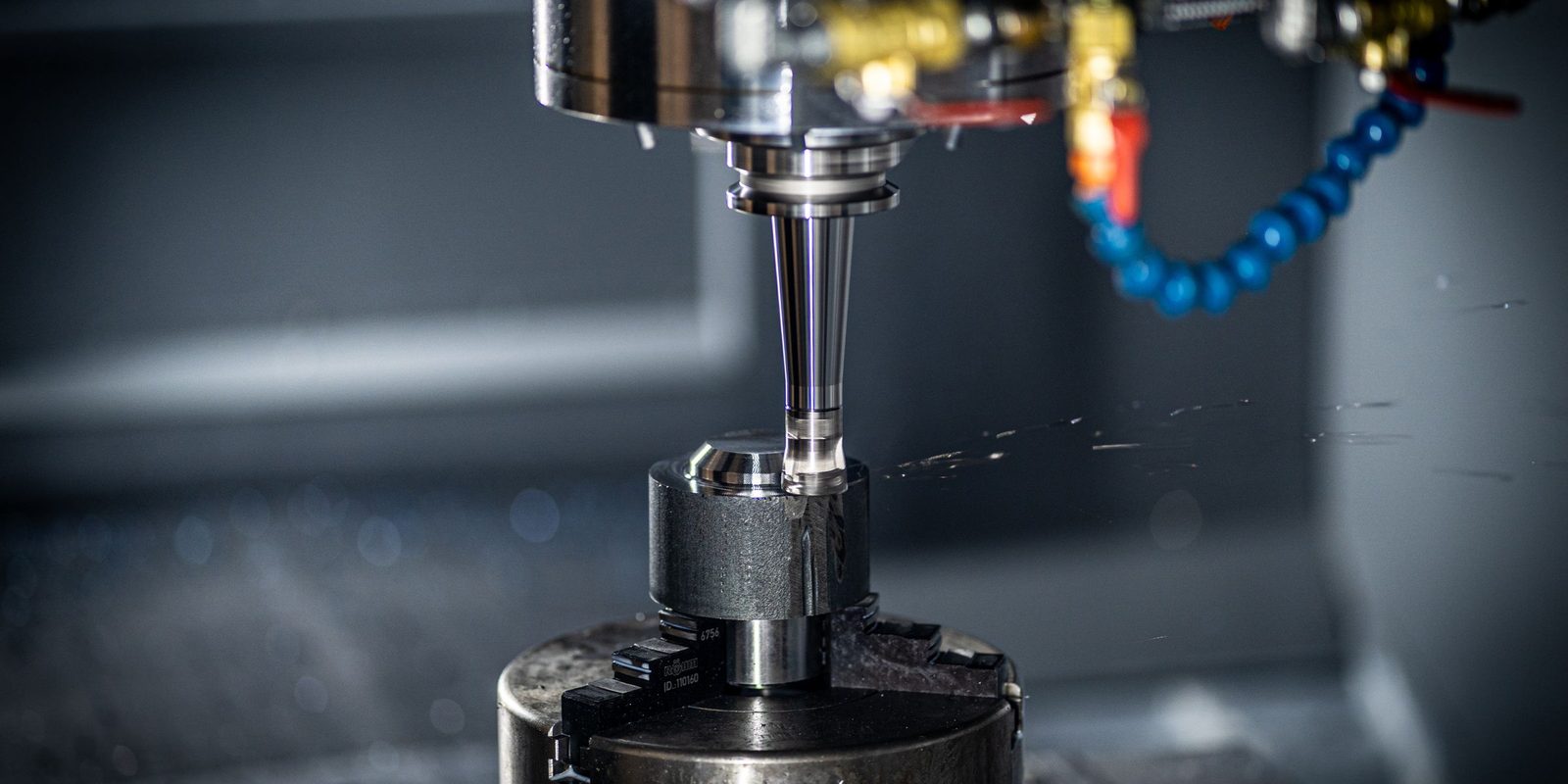 A CNC machining spindle above a part held in a chuck, with a blue coolant hose beside it