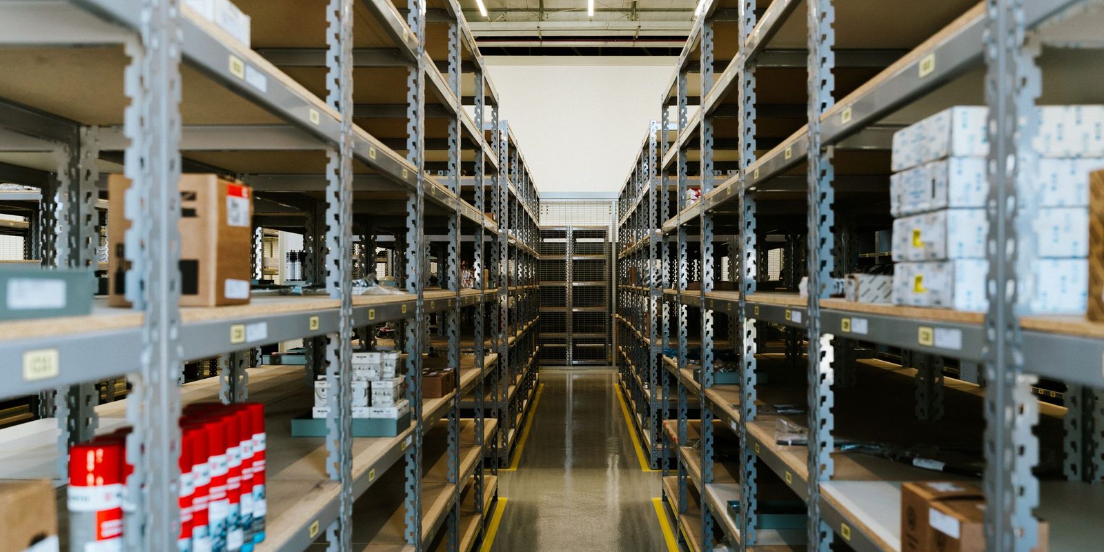 A long aisle of grey steel shelving holding boxed parts, with a polished floor edged by yellow lines