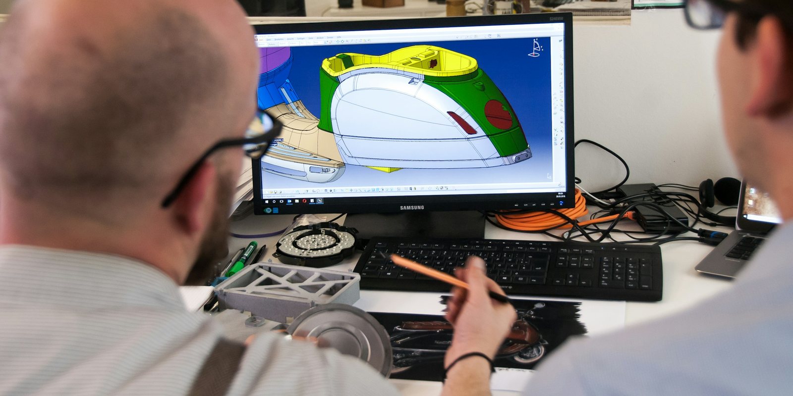 Two engineers at a desk review a colour-coded 3D CAD model of a vehicle body panel on a monitor, with prototype parts beside the keyboard