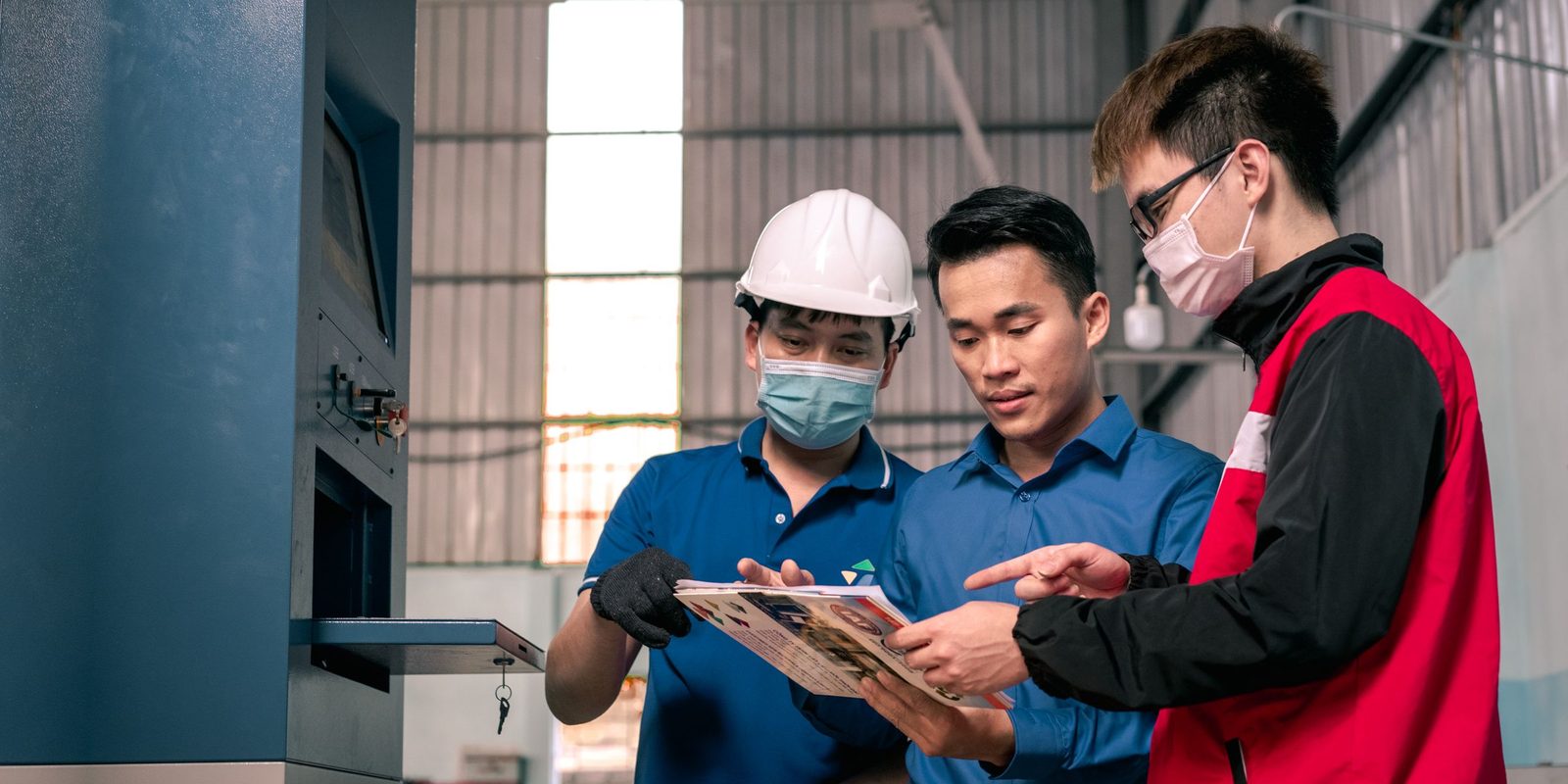 Three colleagues, one in a white hard hat, read a printed sheet together beside a blue machine in a factory