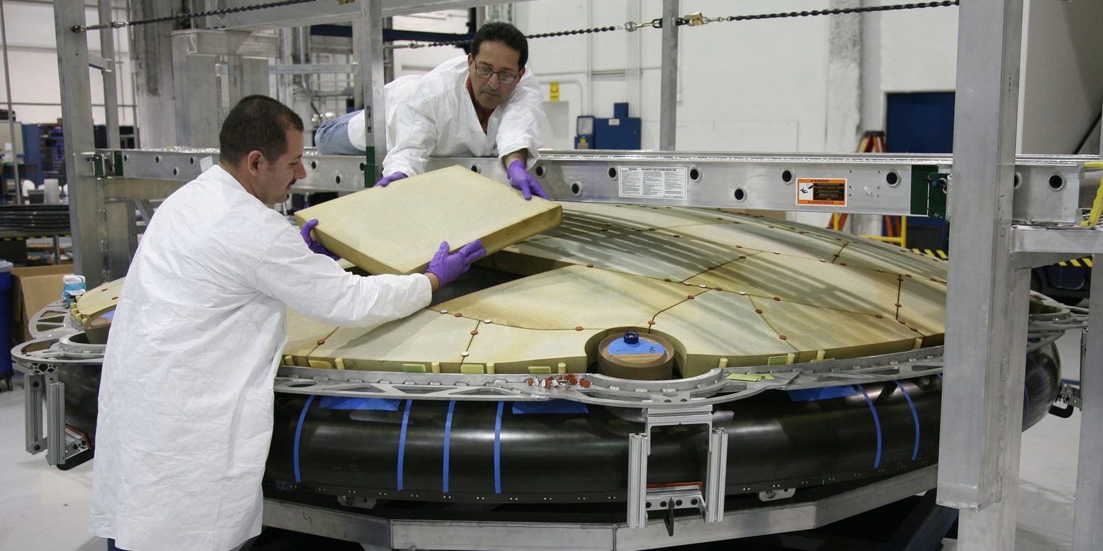 Two technicians in white coats and purple gloves fit panels onto a large round assembly in a bright, clean hall