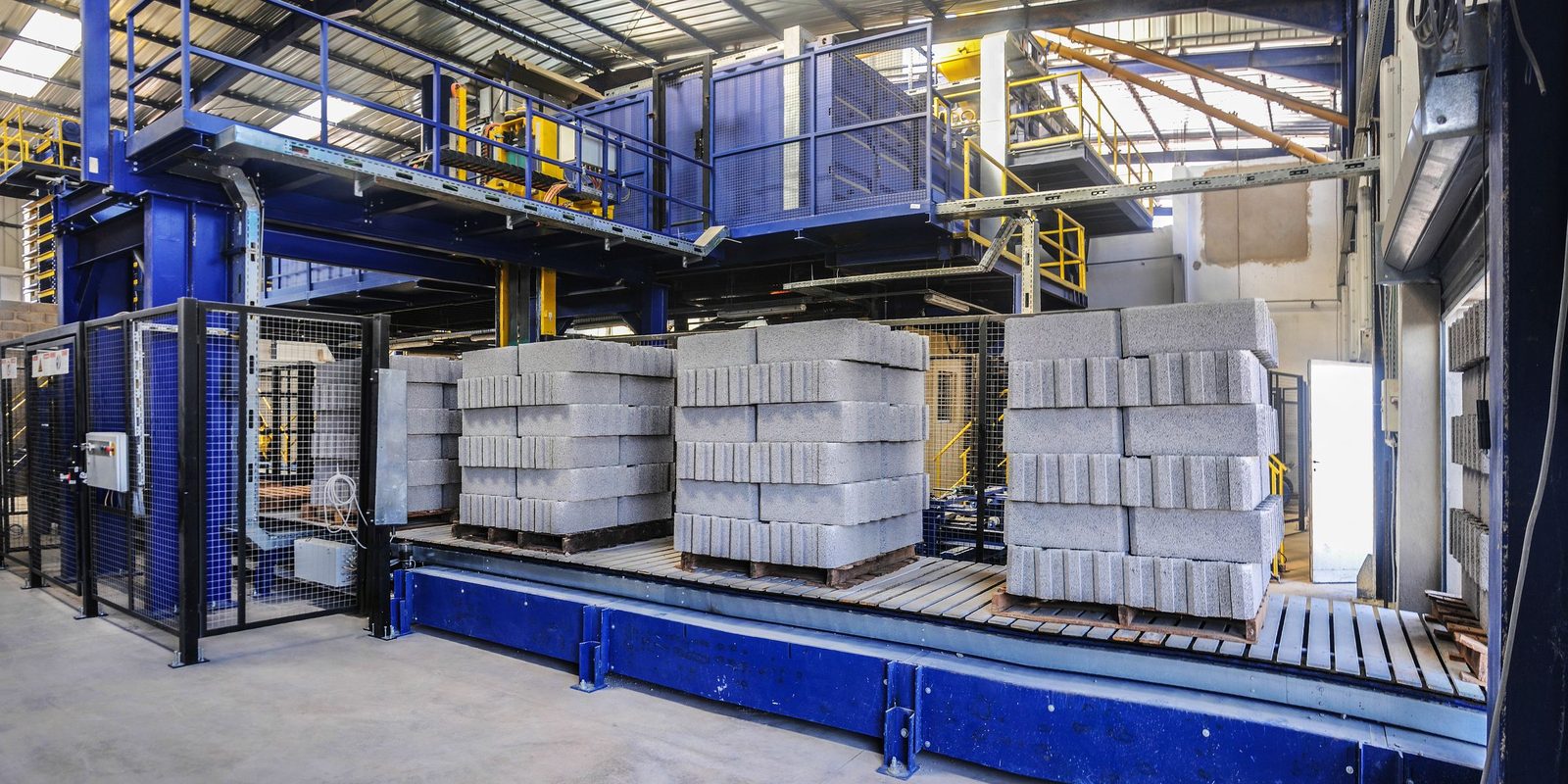 Pallets of concrete blocks wait on a roller conveyor under a blue steel platform in a bright plant