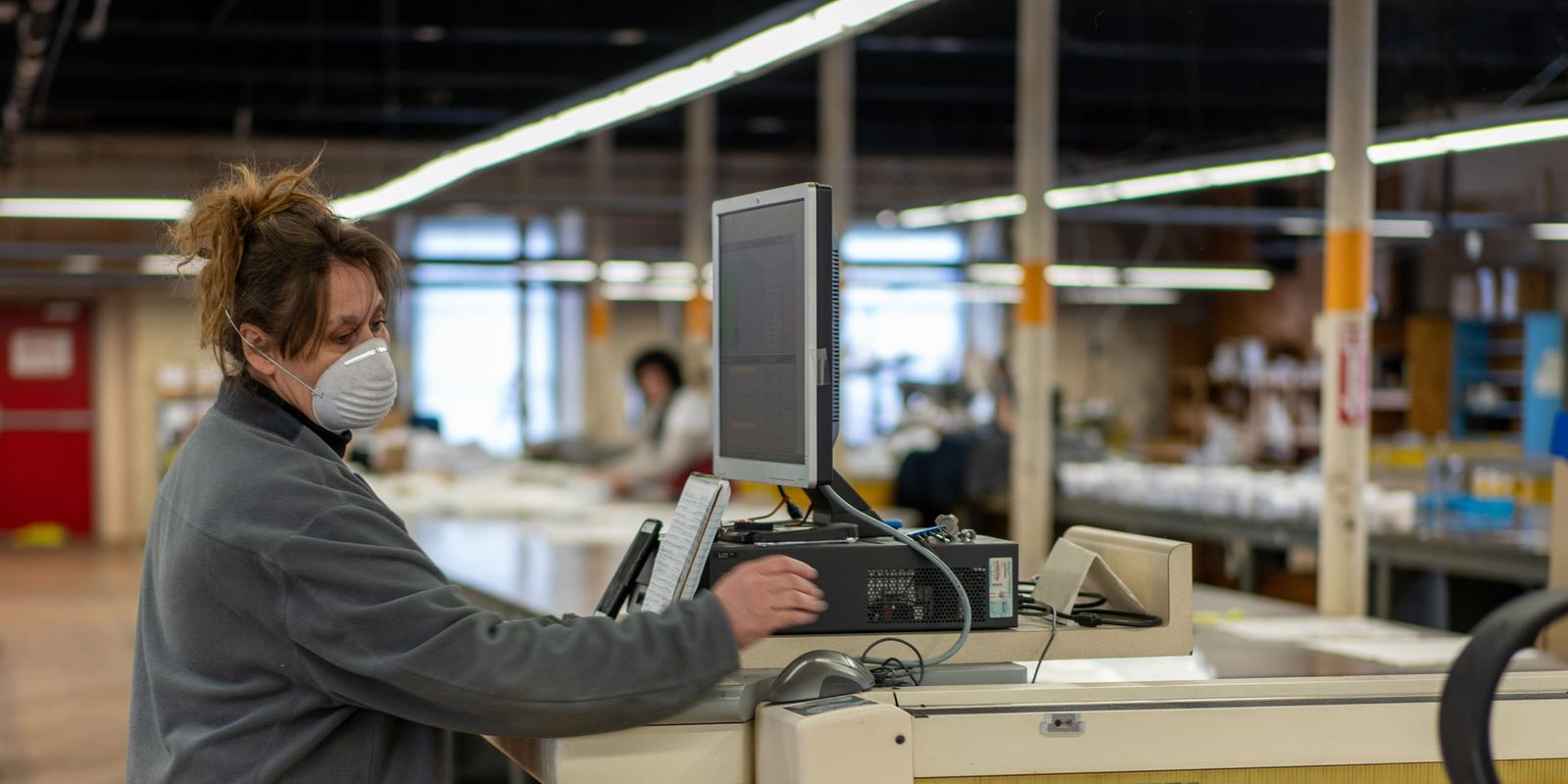 A worker in a face mask uses the computer screen of a cutting table in a bright factory hall
