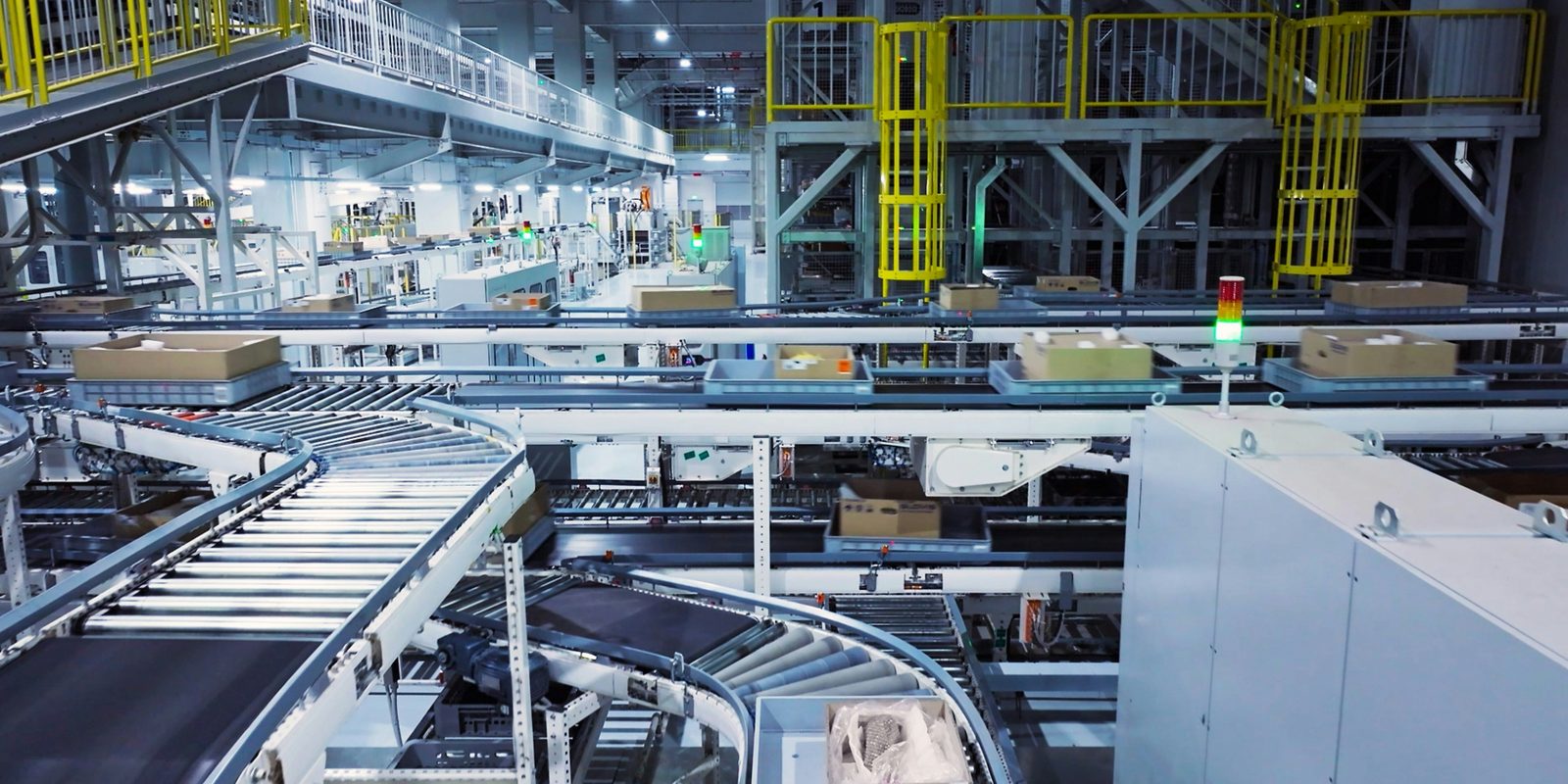 Curved roller conveyors carry totes and boxes through a bright automated production hall with yellow access ladders