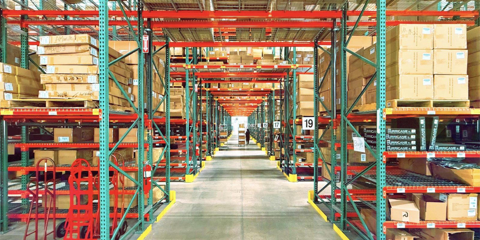 A long aisle of orange and green pallet racking stacked with cartons in a bright distribution warehouse, with a worker and a cart at the far end