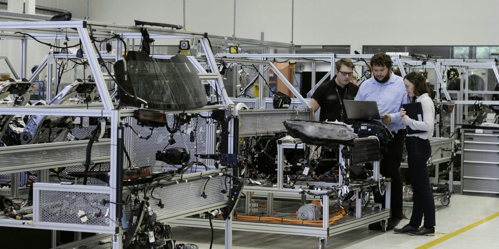 Three engineers review a laptop beside vehicle test rigs on a bright, clean plant floor
