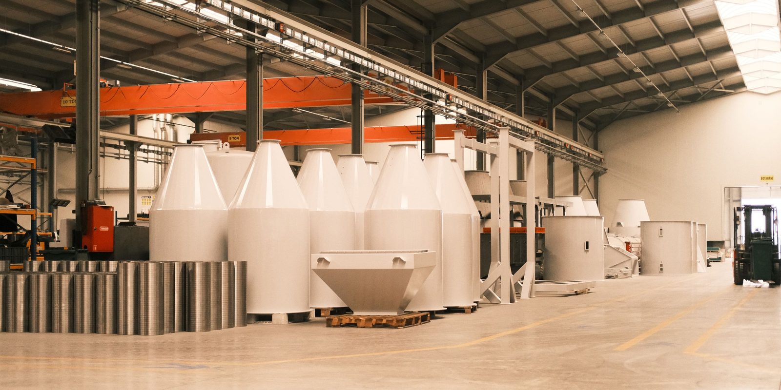 Finished white steel hoppers stand on pallets under orange overhead cranes in a clean, brightly lit fabrication plant