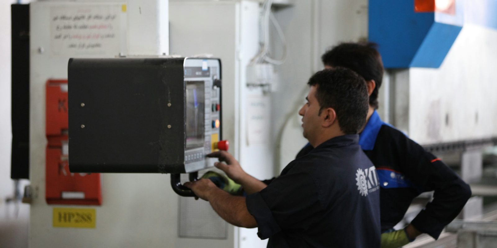 Two operators work at the control pendant of a large CNC machine in a bright fabrication shop