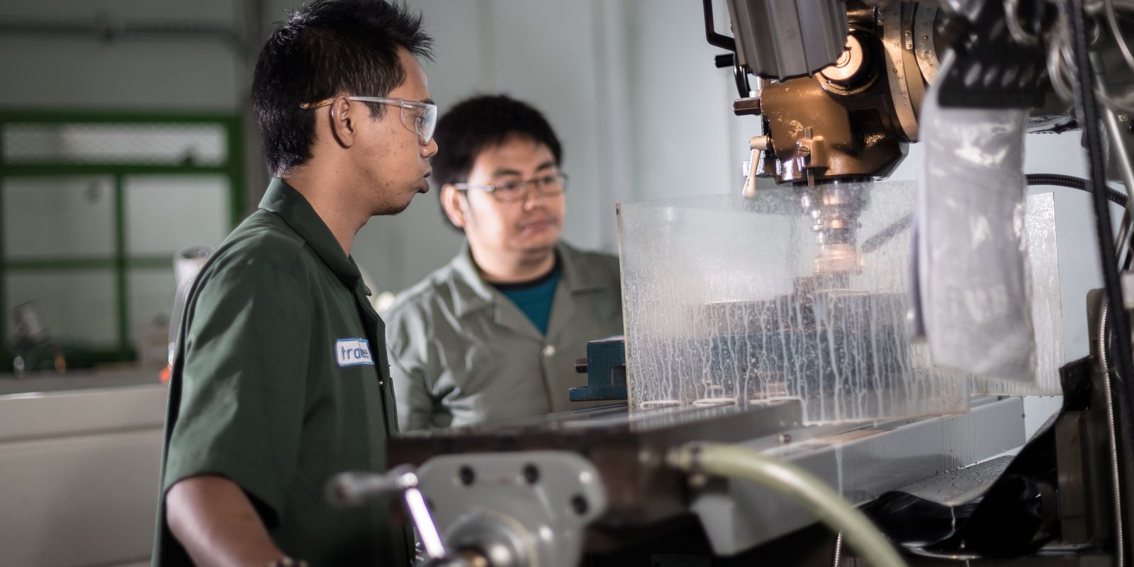 Two technicians watch a CNC milling machine cut a part behind a clear splash guard streaked with coolant, in a bright workshop