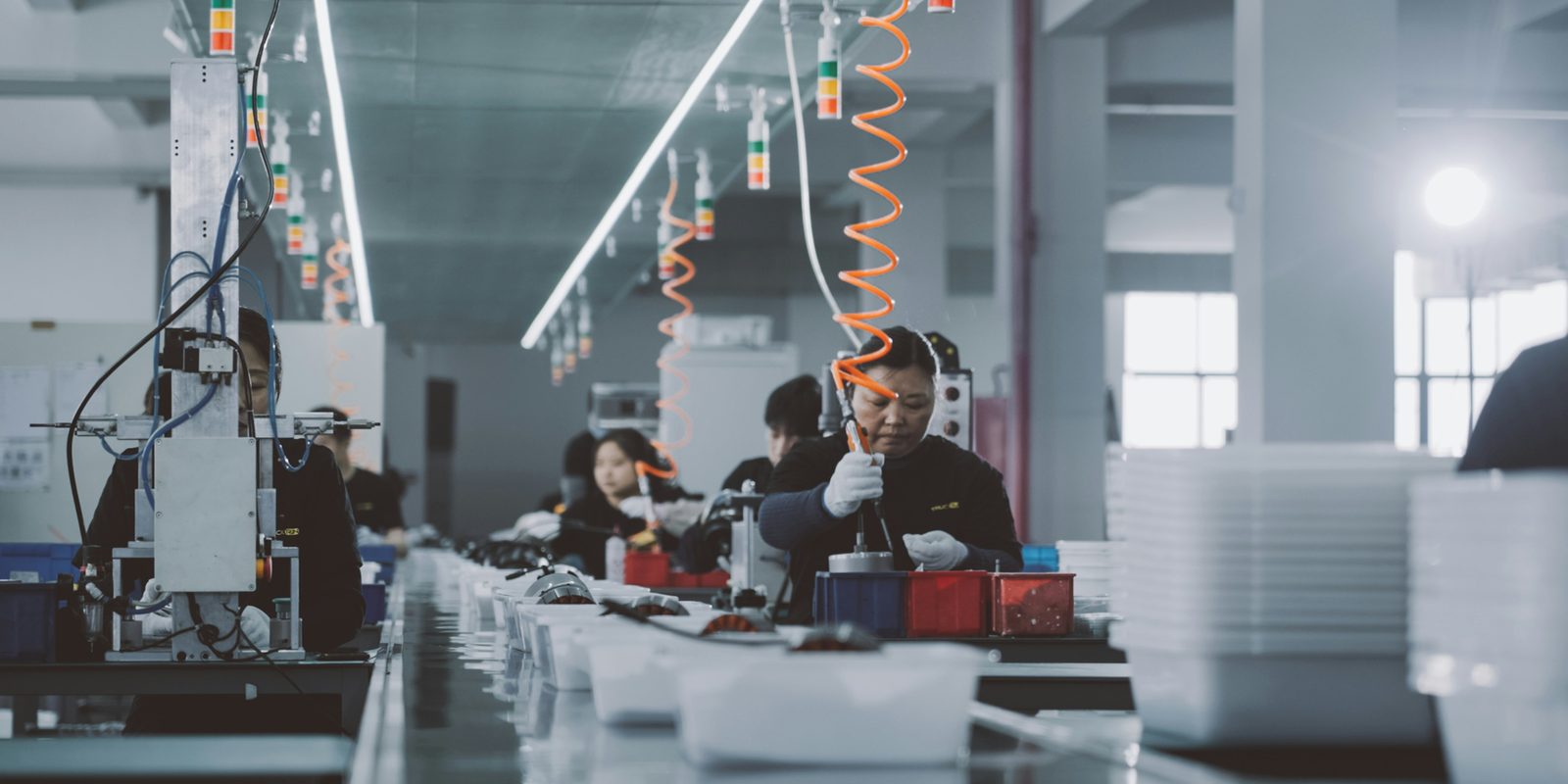 Workers assemble parts at stations along a bright assembly line, with signal lights and air hoses hanging over each station