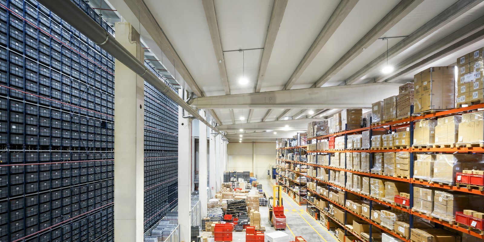 A bright distribution warehouse with a tall wall of automated storage bins on the left and pallet racks of cartons on the right