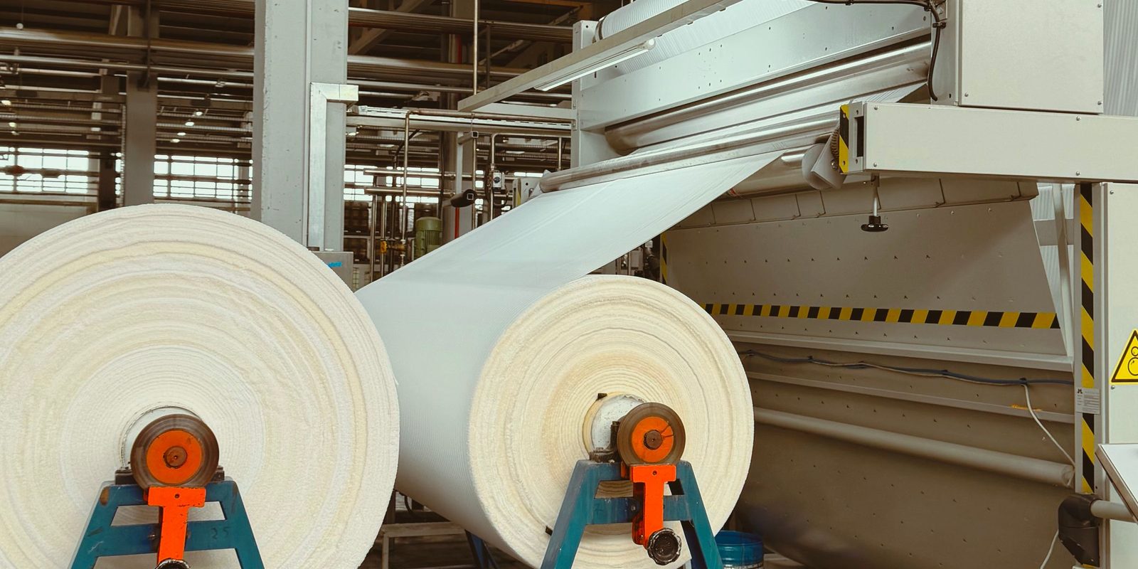 Two large rolls of white fabric on blue stands feed a textile machine in a bright production hall