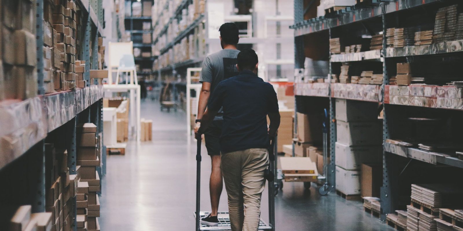 Two men push a cart down a long warehouse aisle lined with tall shelving