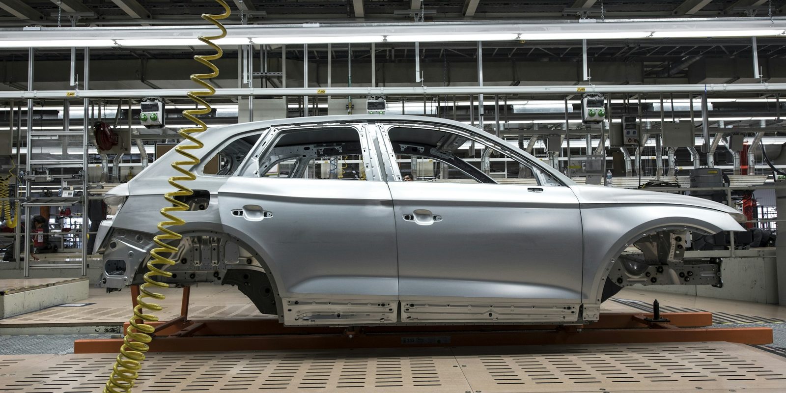 An unpainted car body rides on a carrier through a bright automotive assembly hall