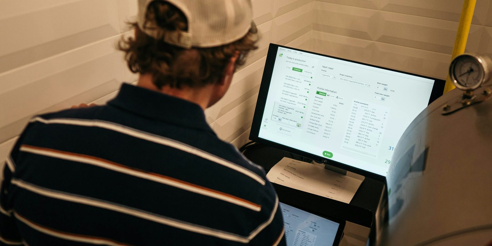 A worker in a cap reads a production screen that lists the day&rsquo;s batches, beside a coffee roasting machine