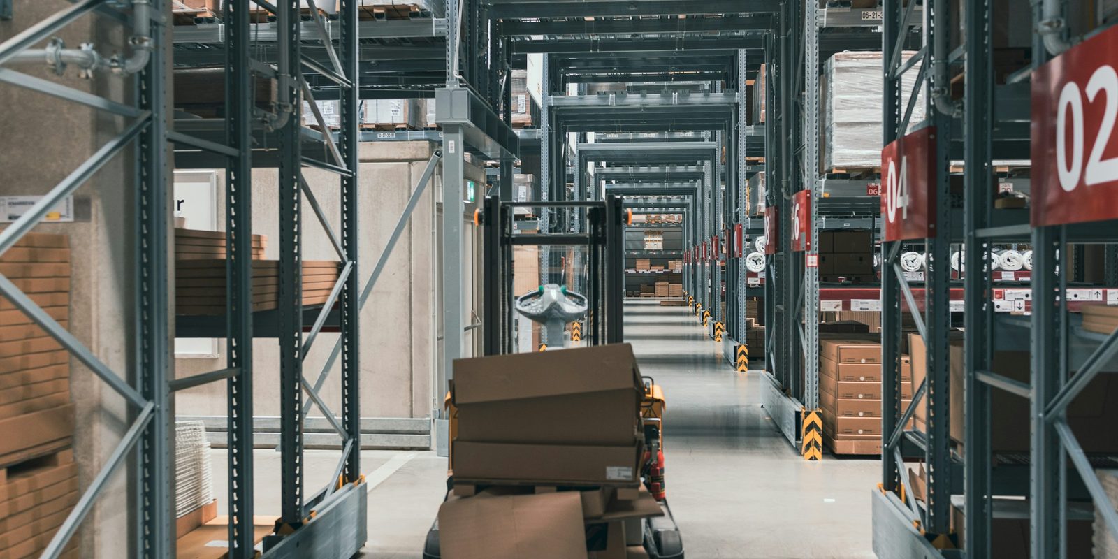 A long warehouse aisle between steel racks with red aisle numbers, and a pallet truck carrying cartons in the foreground