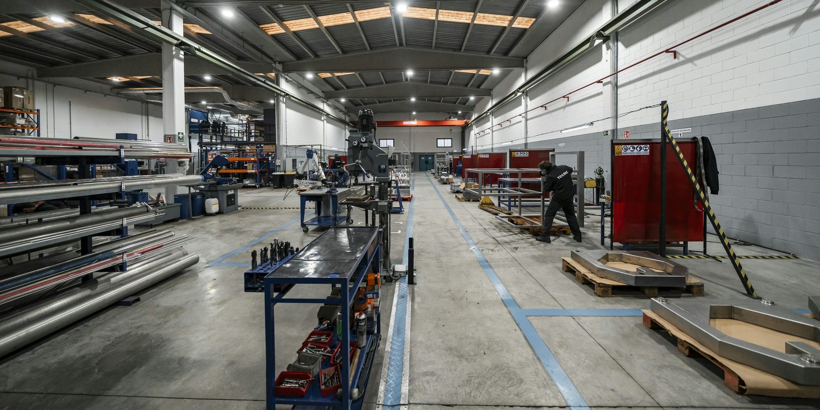 A clean metal fabrication plant with tube racks on the left, workbenches along a marked aisle and a welder at a frame on the right