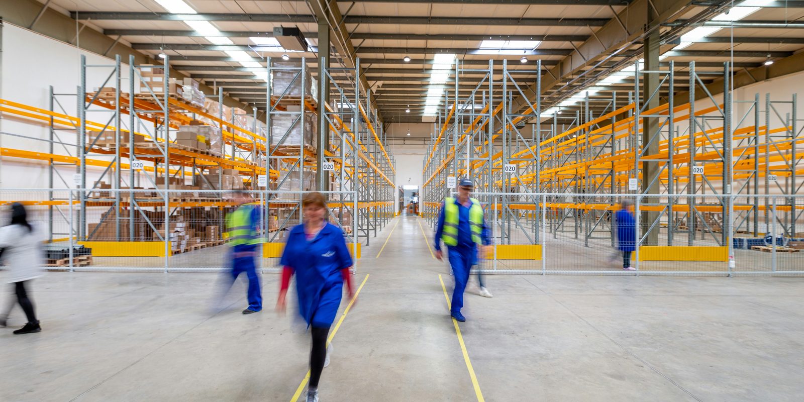Staff in blue workwear walk the main aisle of a bright distribution warehouse lined with pallet racking