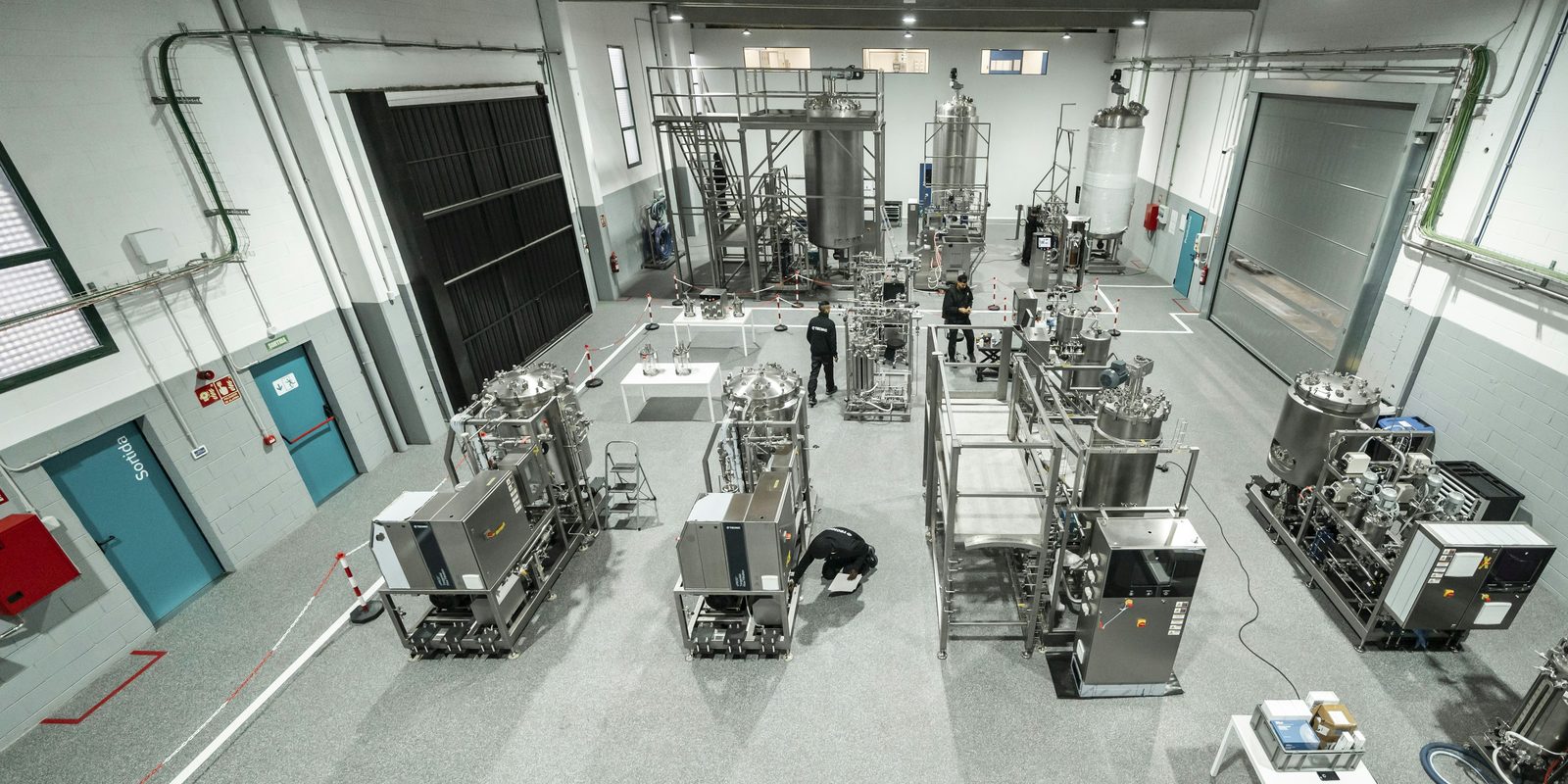 Overhead view of a bright production hall with stainless process skids laid out in rows and two technicians at work