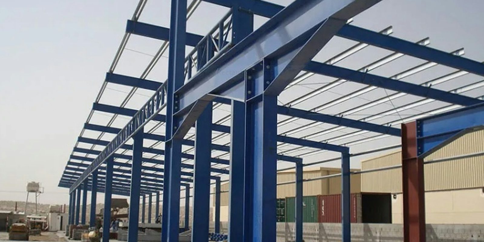 The blue steel frame of a large industrial building going up, with columns and roof beams in place under a clear sky