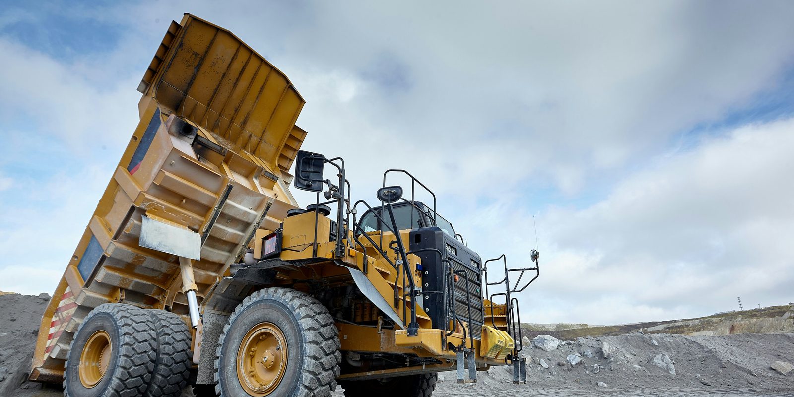 A yellow mining haul truck raises its dump body under a bright, cloudy sky