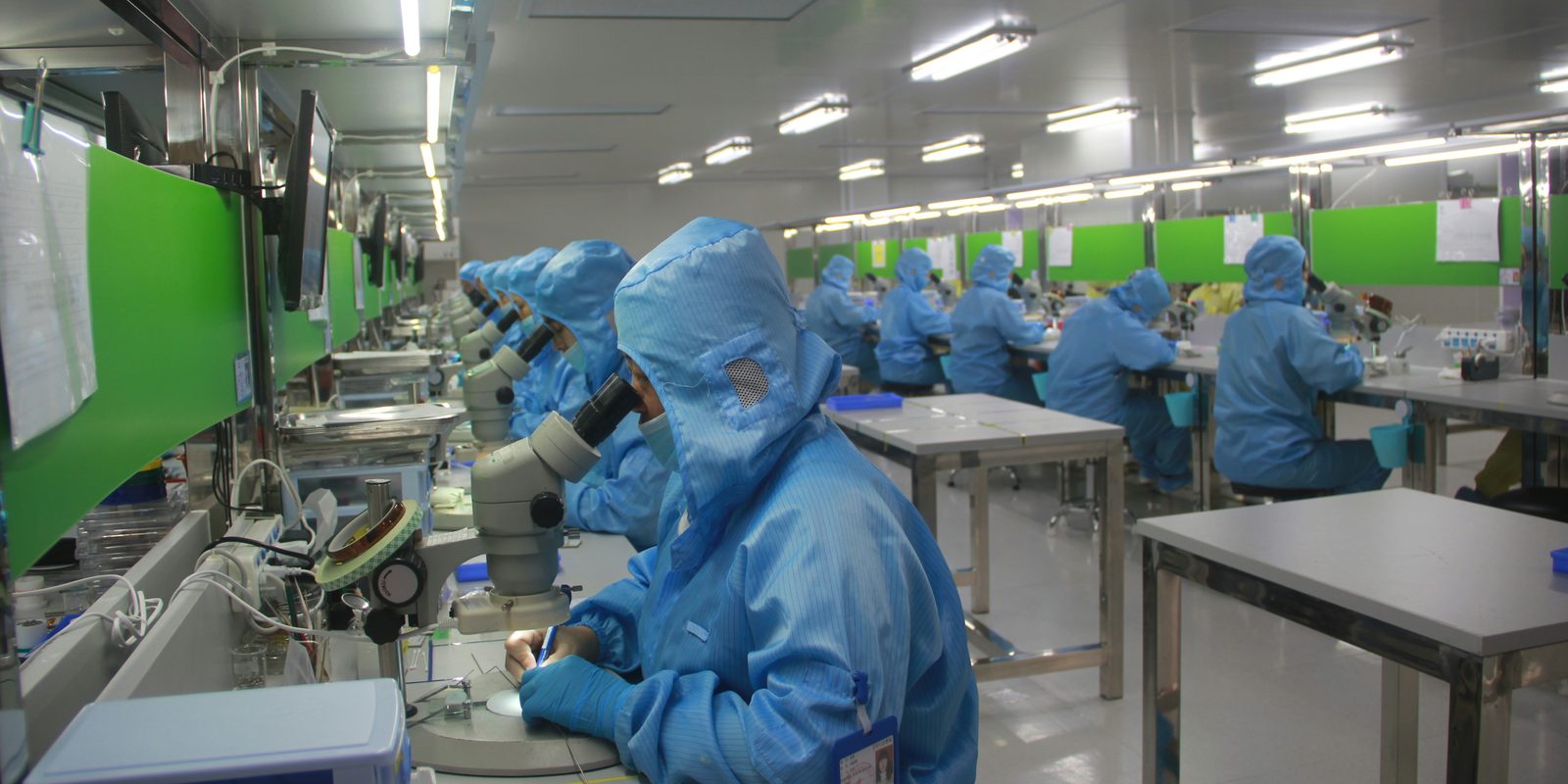 Rows of technicians in blue cleanroom suits assemble electronics at microscope benches in a bright plant