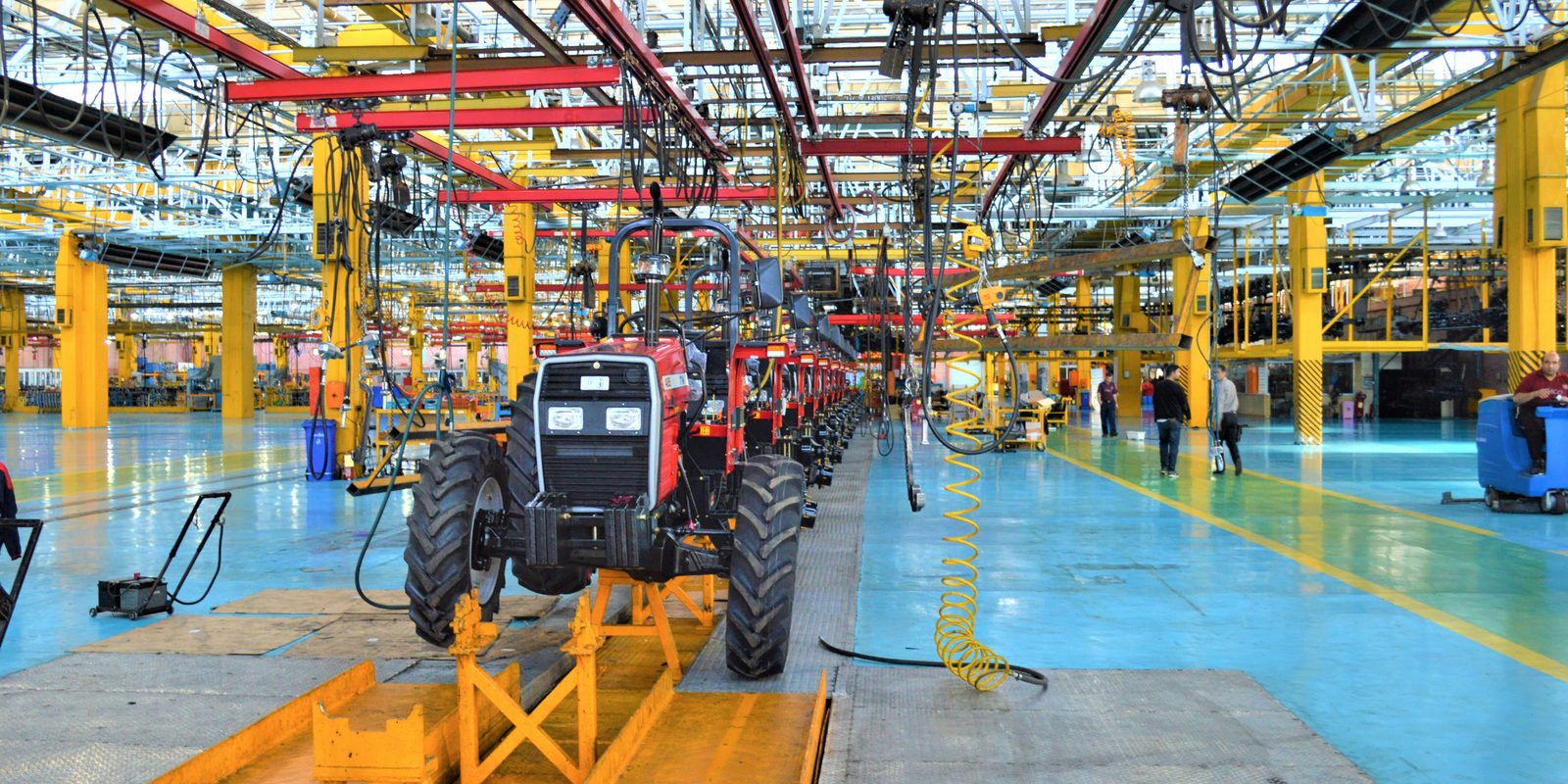 Red tractors lined up on an assembly line under yellow and red overhead rails in a bright plant with a blue floor