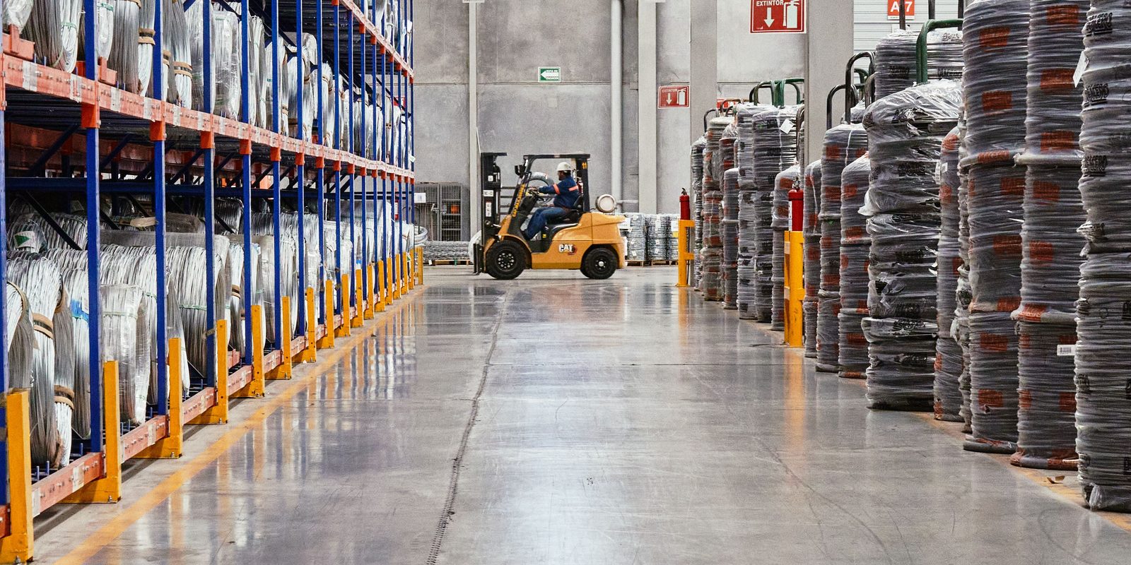 A bright, orderly warehouse aisle with racks of stock on both sides and a forklift at the far end