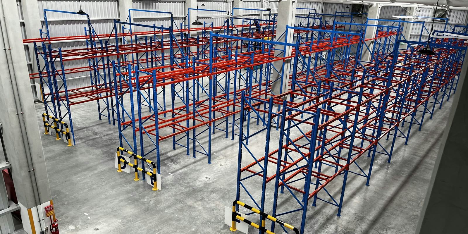 Rows of empty blue and red pallet racking on a clean concrete floor, with yellow and black guard rails at the aisle ends