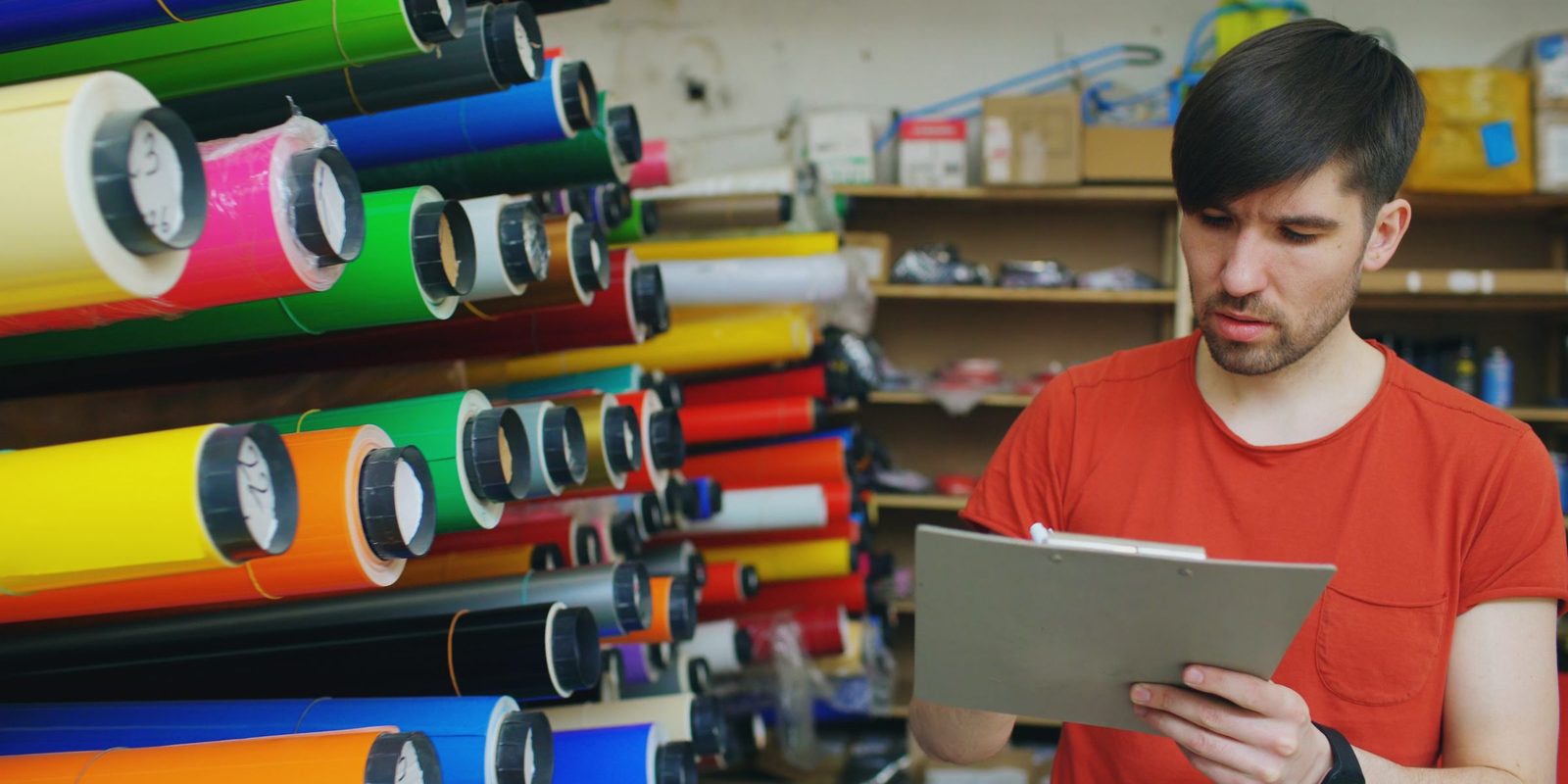 A man in a red T-shirt reads a clipboard beside racks of coloured vinyl rolls, the roll ends marked with handwritten numbers