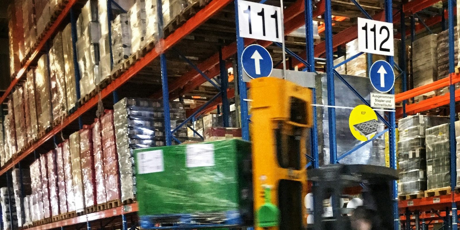 A reach truck carries a wrapped pallet down an aisle of tall pallet racks marked with the location numbers 111 and 112