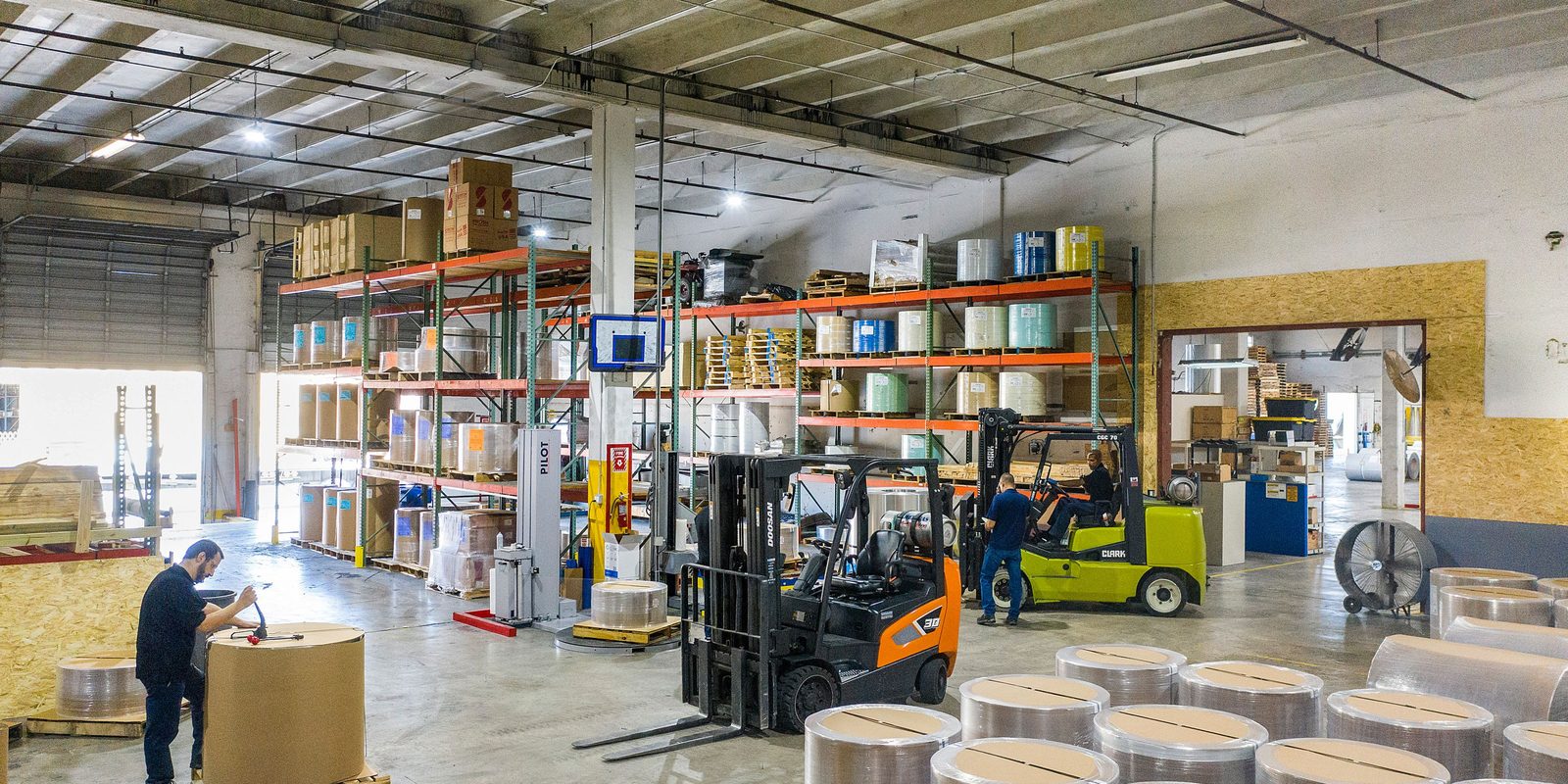Two forklifts work between pallet racks of film rolls and boxes in a bright distribution warehouse while a worker prepares a roll in the foreground