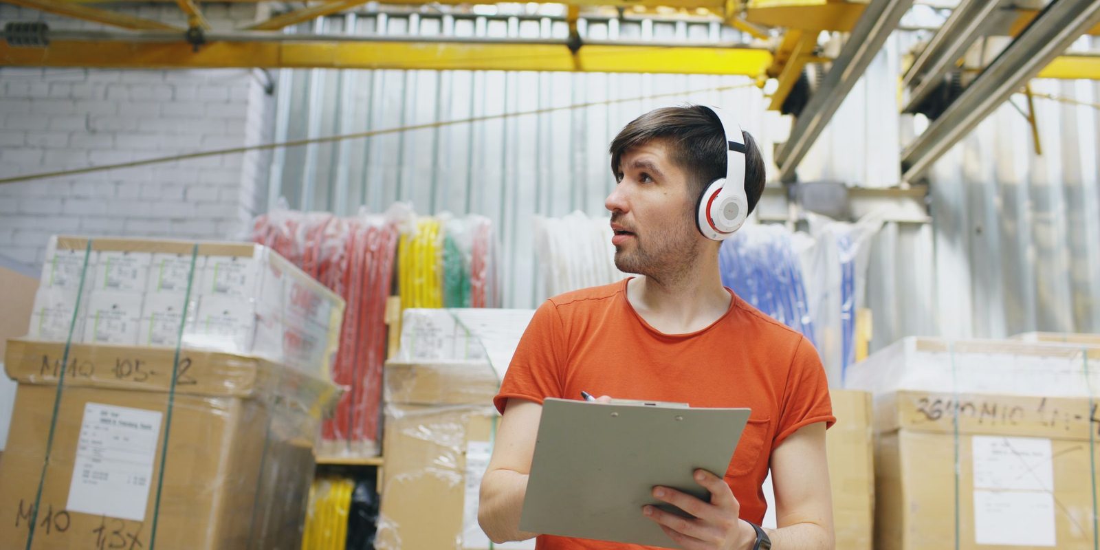 A warehouse worker in an orange T-shirt and headphones holds a clipboard beside taped cartons, with bundles of red, yellow and blue stock behind him