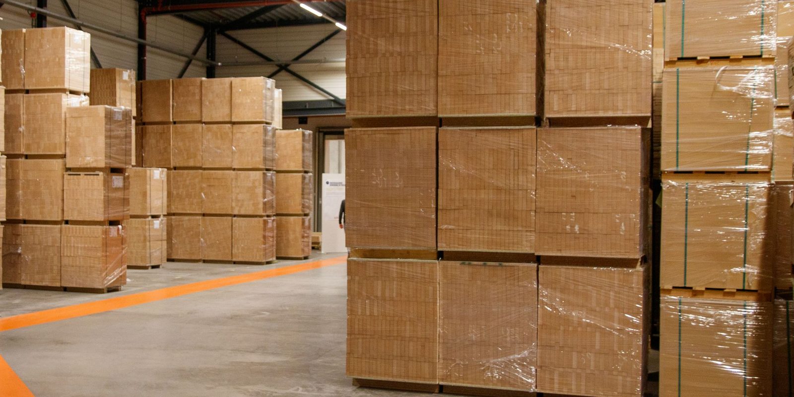 Shrink-wrapped pallets of wooden blocks stacked in a warehouse beside an orange floor lane