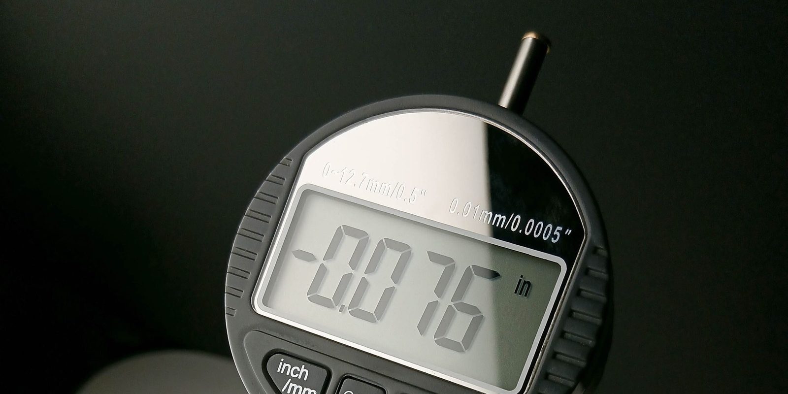 Close-up of a digital measuring gauge whose display reads minus 0.076 inches, against a dark background