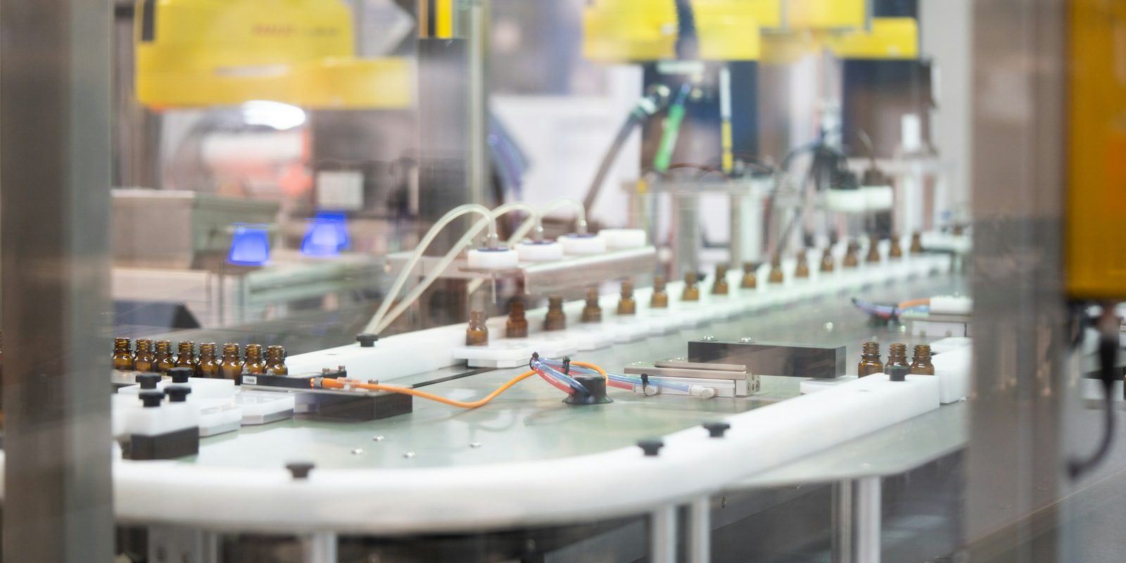 Small amber glass vials move along a white track on an automated filling line, with yellow robots out of focus above