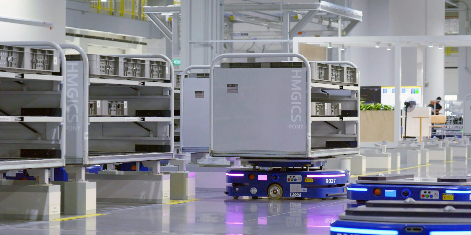 Blue mobile robots carry racks of parts bins across the polished floor of a bright, automated plant