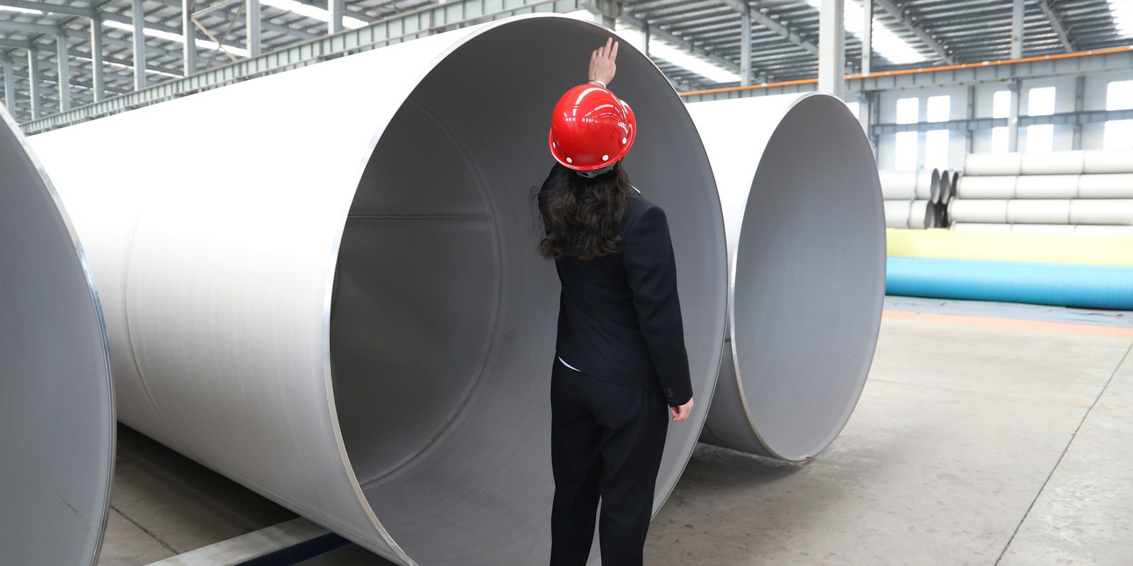 An inspector in a red hard hat checks the inside of a large stainless steel pipe in a bright factory