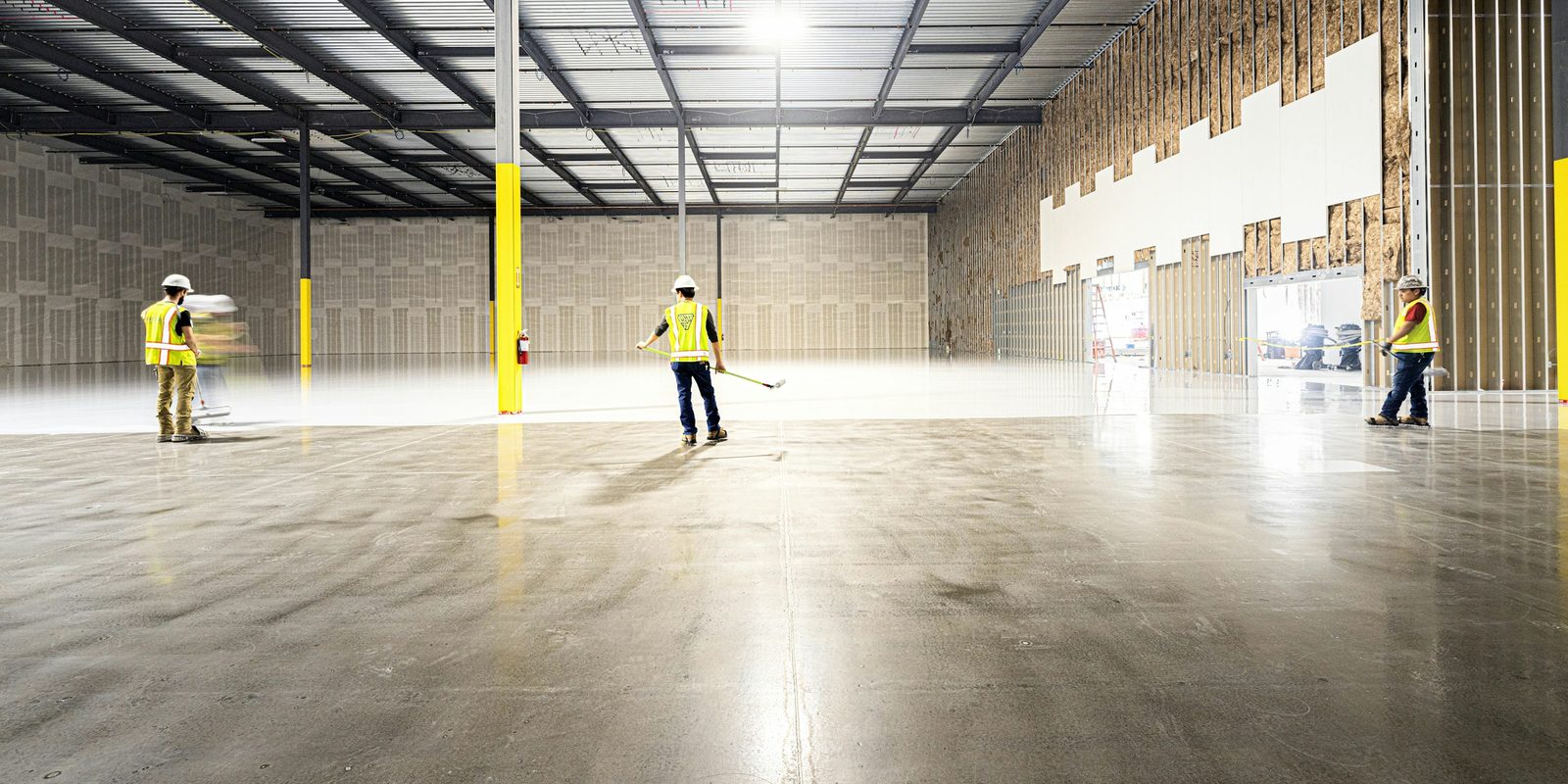 Three workers in high-visibility vests and hard hats finish the polished floor of a large, bright new warehouse