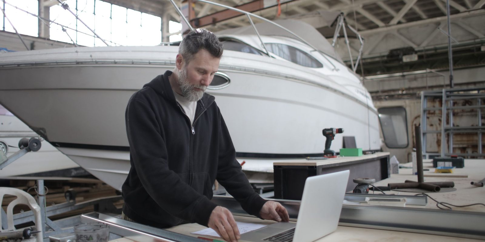 A man with a grey beard works on a laptop at a workbench in a bright boat workshop, with a white motorboat behind him