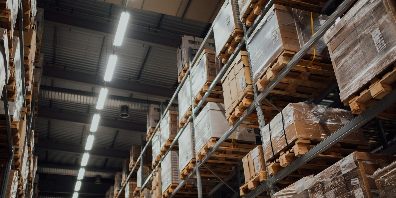 Pallets of shrink-wrapped cardboard boxes stacked on tall steel racking under a line of ceiling lights in a warehouse aisle