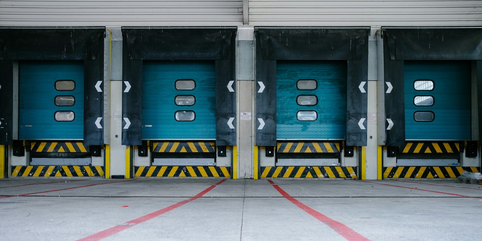 Four loading dock doors with blue roll-up doors, black dock seals and yellow and black safety markings above a concrete apron with red painted lines