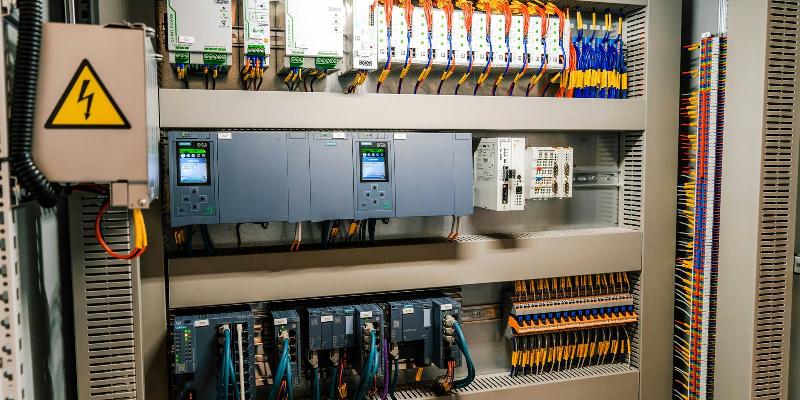 The inside of an industrial control cabinet with PLC modules, breakers, relays and rows of colour-coded terminal blocks