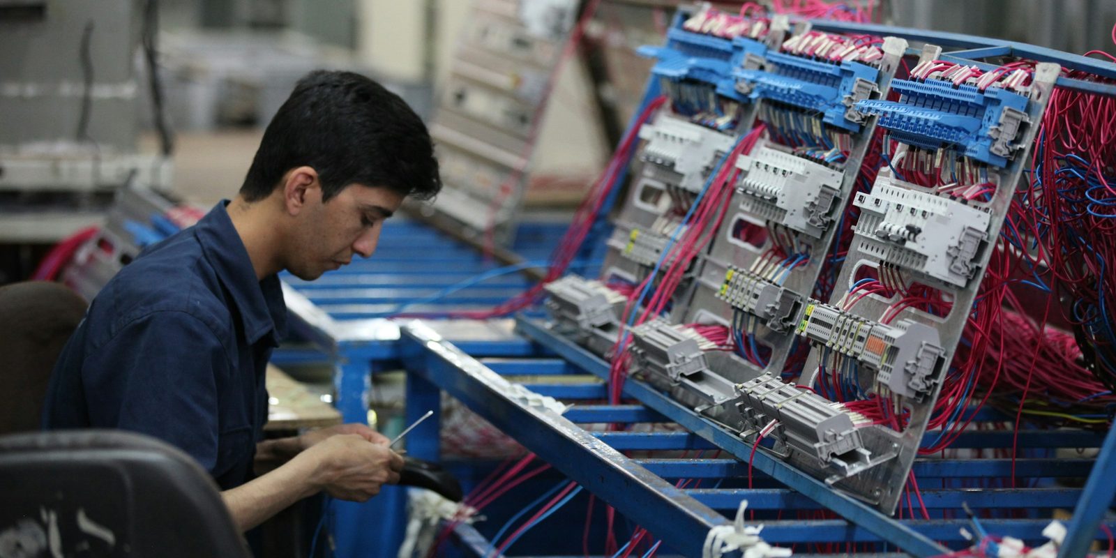 A technician wires breakers and terminal blocks on electrical panels mounted on a blue assembly rack in a bright workshop
