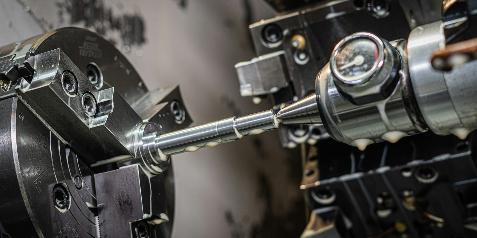 A steel shaft turning on a CNC lathe, held in a three-jaw chuck and supported by a live centre