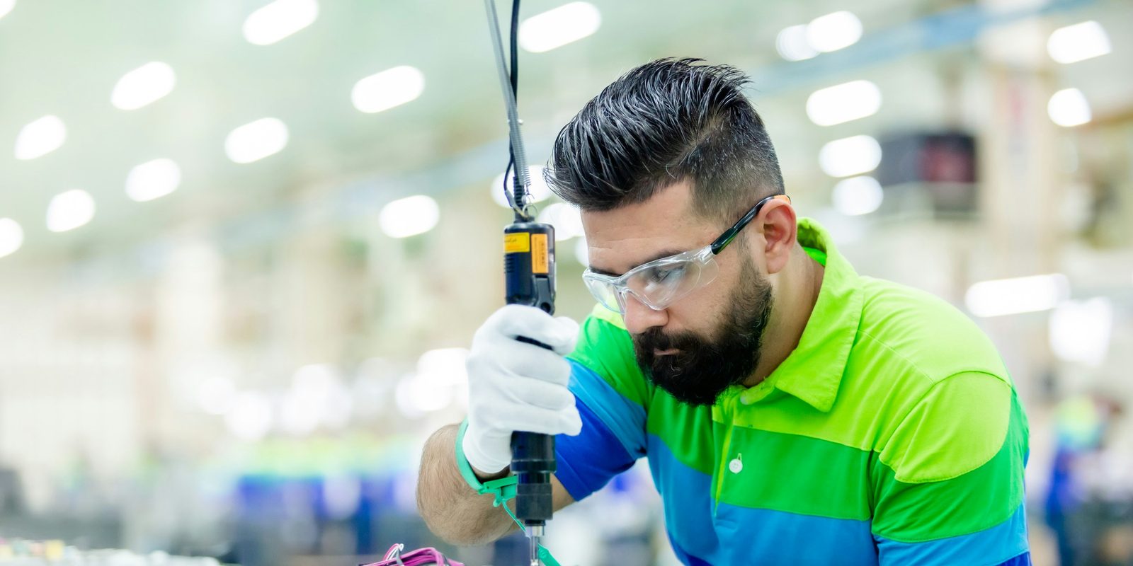 Technician in safety glasses uses a suspended electric screwdriver on an assembly bench in a bright factory