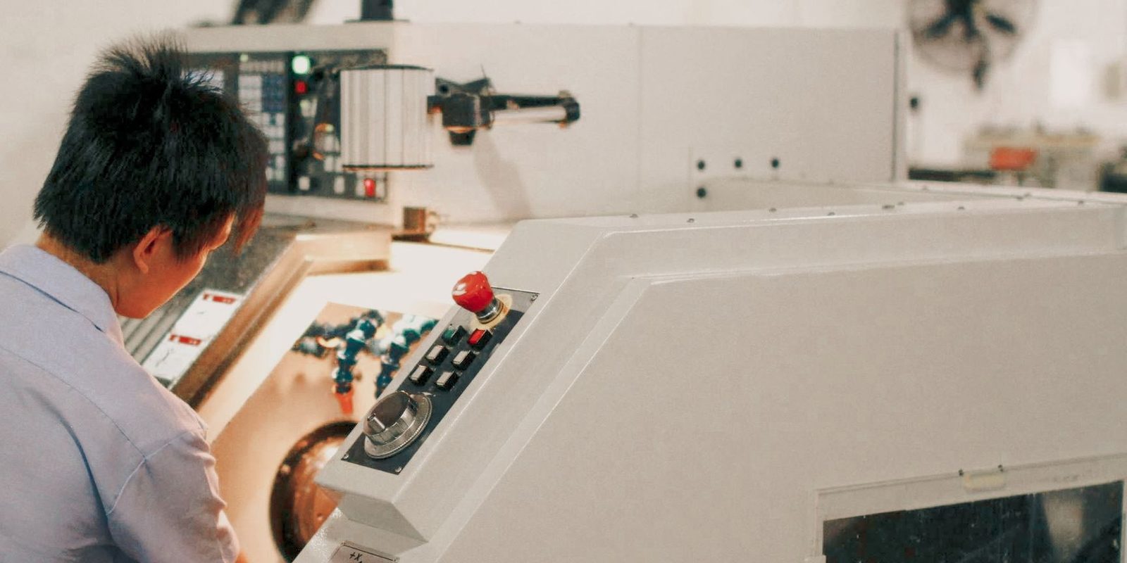 A machinist in a light blue shirt reaches into a white CNC lathe next to the control panel