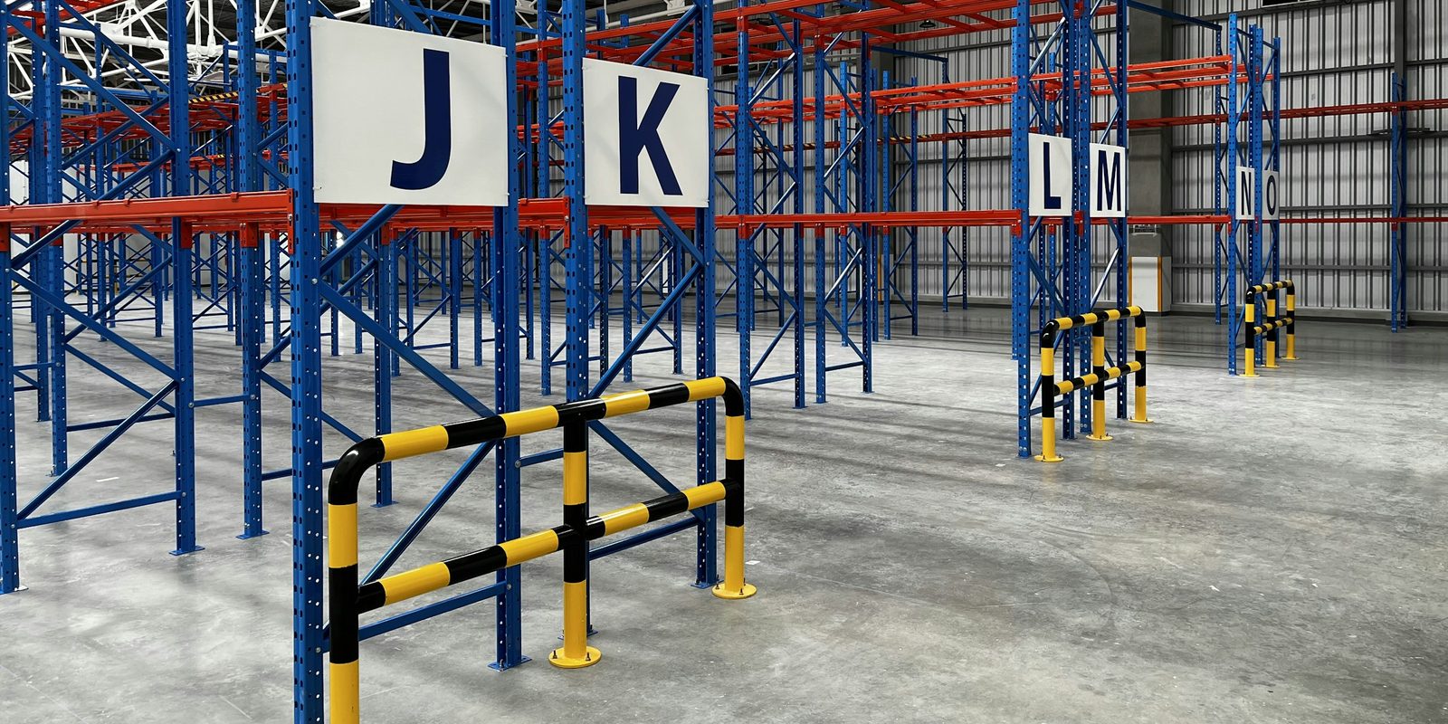 Blue and orange pallet racking with large aisle signs J, K, L, and M in an empty, bright warehouse